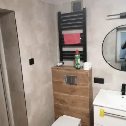 Nowoczesna łazienka z beżowymi płytkami, podwieszaną toaletą z drewnopodobną obudową, czarnym grzejnikiem drabinkowym z ręcznikiem i okrągłym lustrem