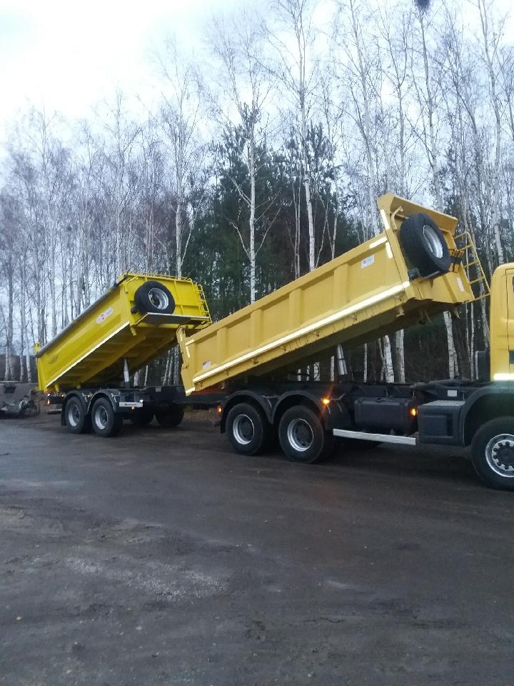 Żółta, dwu-przyczepowa wywrotka z podniesionymi kiprami, na tle drzew bezlistnych, na placu roboczym.