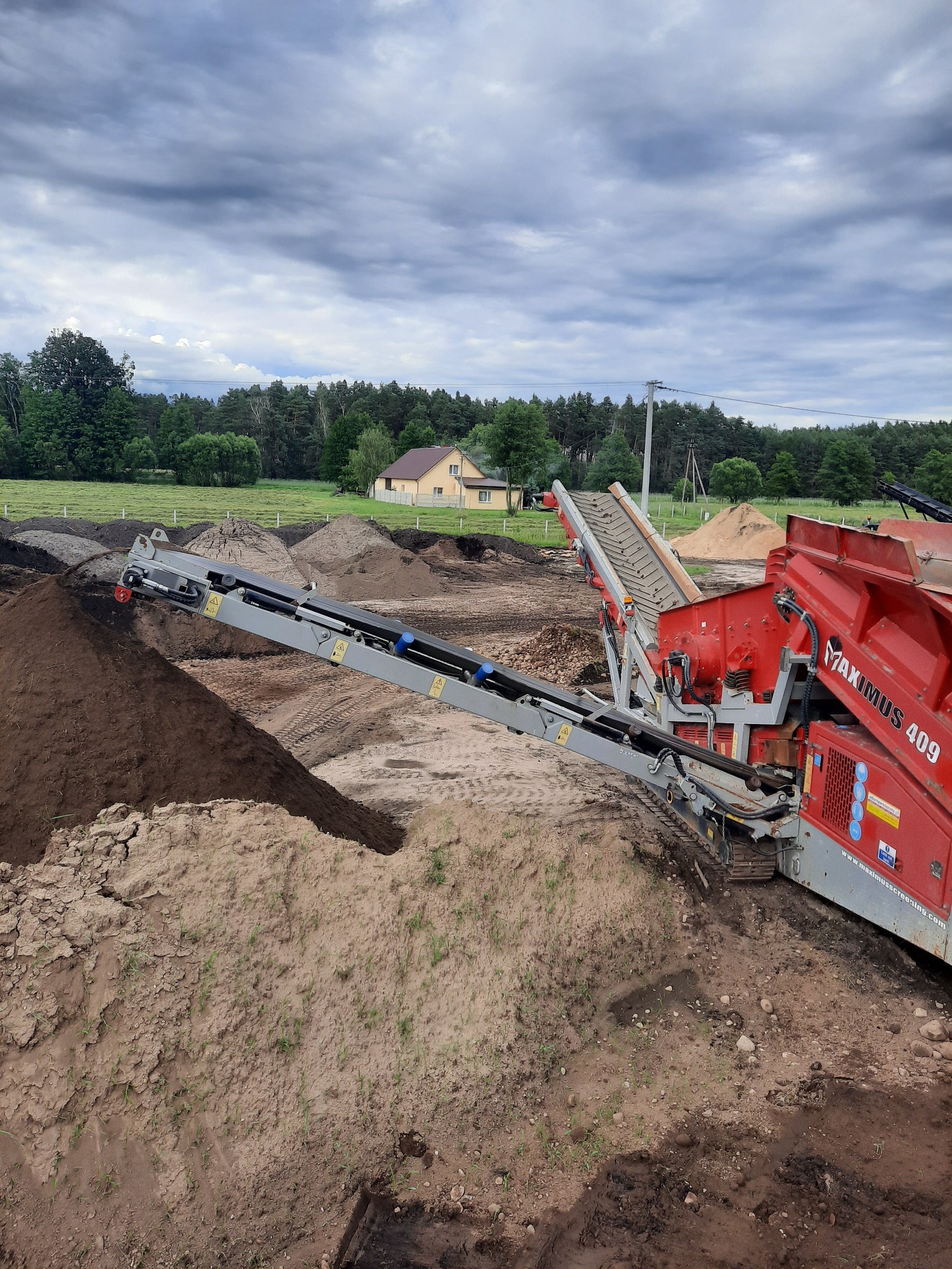 Czerwona przesiewarka mobilna Maximus 409 z wysięgnikiem taśmowym, usypująca ciemną ziemię na pryzmę, w tle widoczne pole, dom i pochmurne niebo.