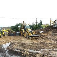 Żółta koparko-ładowarka na błotnistym placu budowy z wykopaną ziemią i widocznymi elementami infrastruktury w tle.