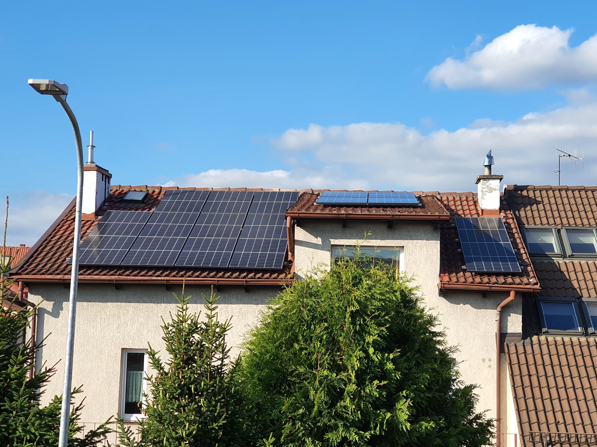Panele słoneczne na dachu dwupiętrowego domu z czerwonej dachówki, widoczne z poziomu ulicy, z lampą uliczną po lewej stronie i zielonymi krzewami na pierwszym planie.