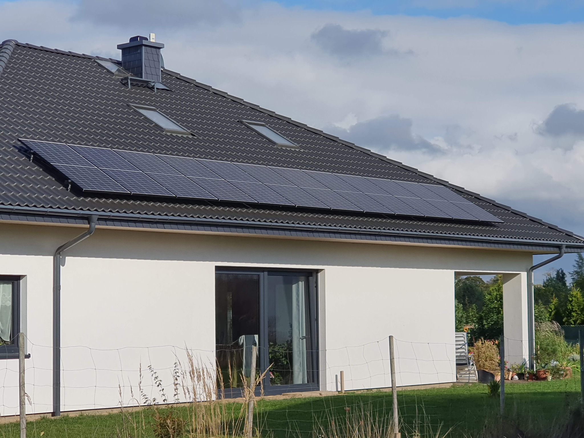 Panele słoneczne na dachu domu jednorodzinnego w Słupsku, widoczne okna dachowe i czarny komin, biała elewacja budynku.