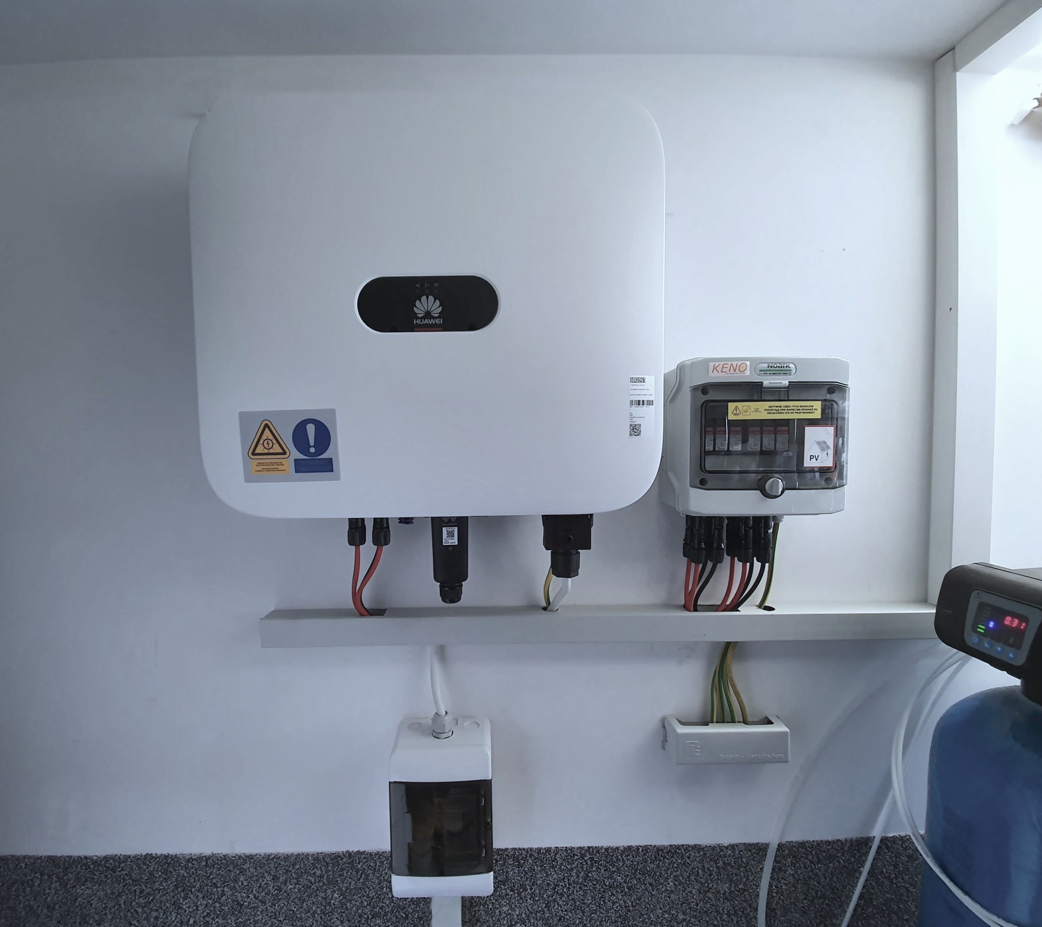 Zamontowany, biały falownik Huawei oraz skrzynka zabezpieczeń KENO z widocznymi przewodami instalacji fotowoltaicznej na jasnej ścianie.