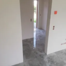 Wnętrze w trakcie prac wykończeniowych, widoczne gładkie, białe ściany, szara podłoga z płytek imitujących kamień, otwór drzwiowy z widokiem na zieleń na zewnątrz, oraz zabezpieczone folią otwory...