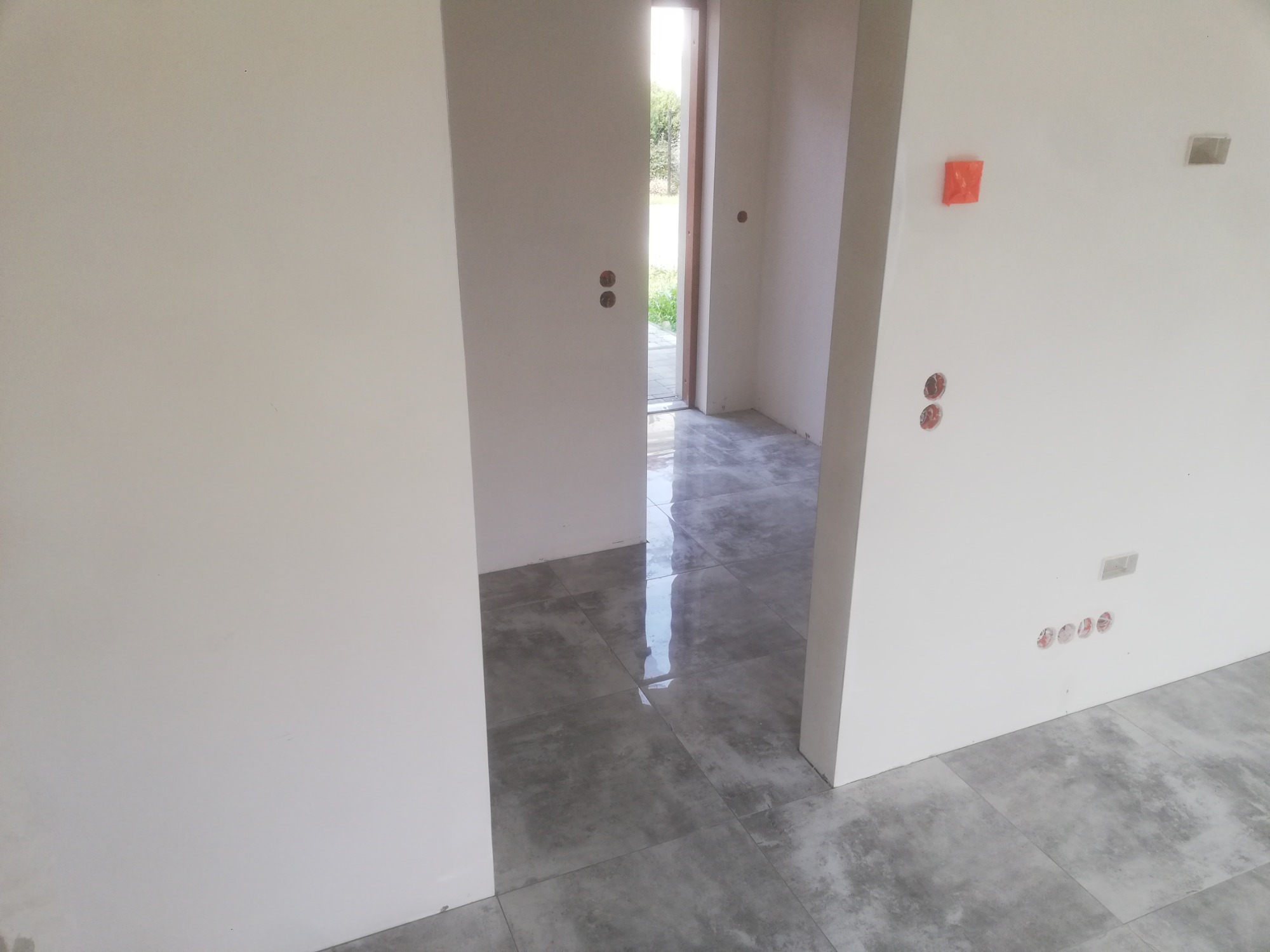 Wnętrze w trakcie prac wykończeniowych, widoczne gładkie, białe ściany, szara podłoga z płytek imitujących kamień, otwór drzwiowy z widokiem na zieleń na zewnątrz, oraz zabezpieczone folią otwory...