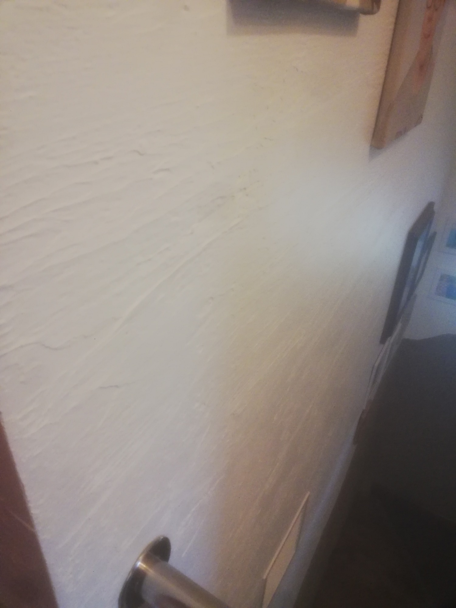 Teksturowana, biała ściana po świeżym malowaniu, widoczny fragment metalowego uchwytu i dekoracje ścienne w tle.