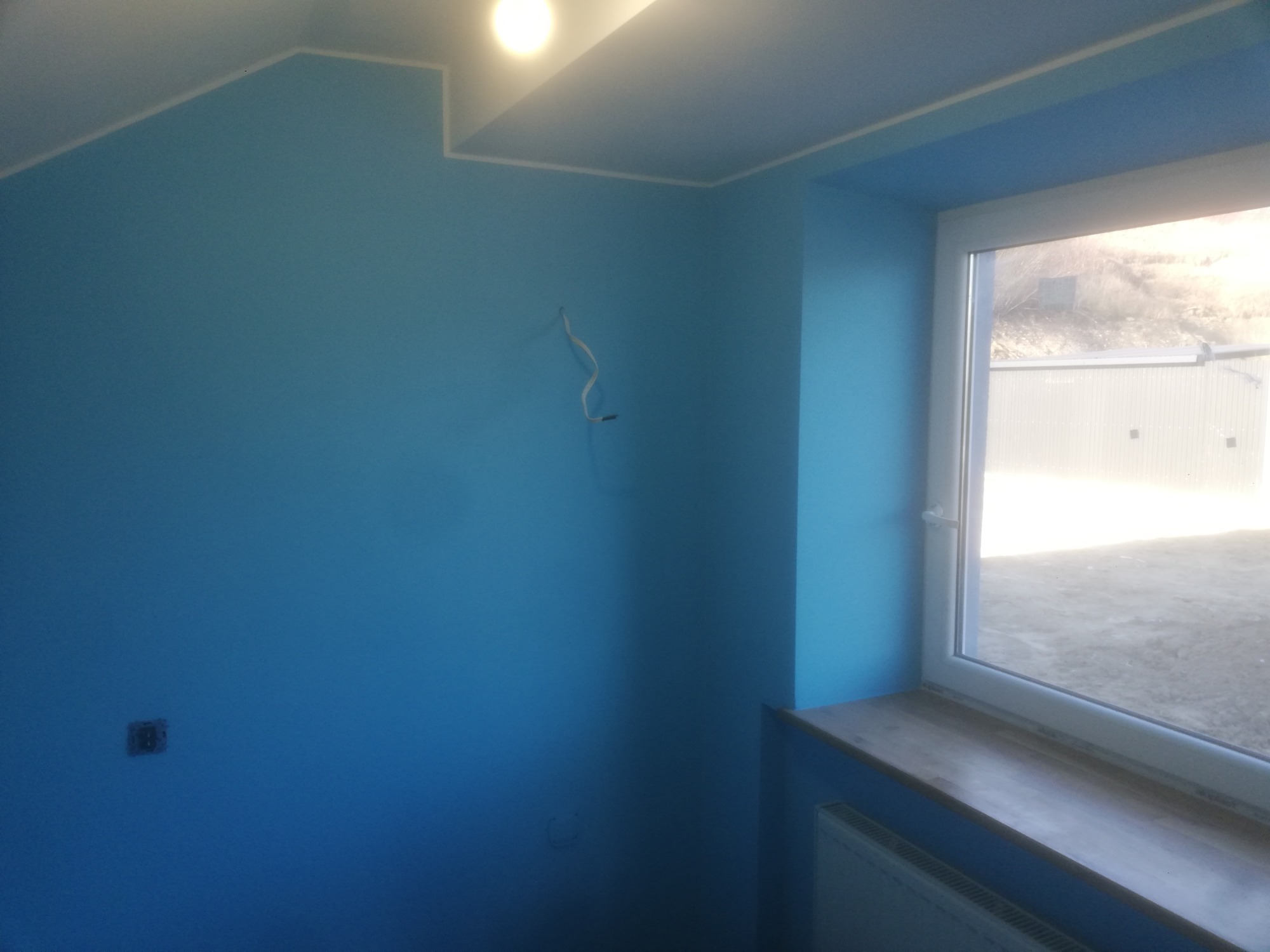 Pomieszczenie po malowaniu na niebiesko, widoczne wystające kable, gniazdko elektryczne i parapet okienny z widokiem na zewnątrz.