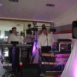 Zespół muzyczny w trakcie występu na sali bankietowej, widoczne instrumenty, nagłośnienie, oświetlenie i baner reklamowy z numerami telefonów.