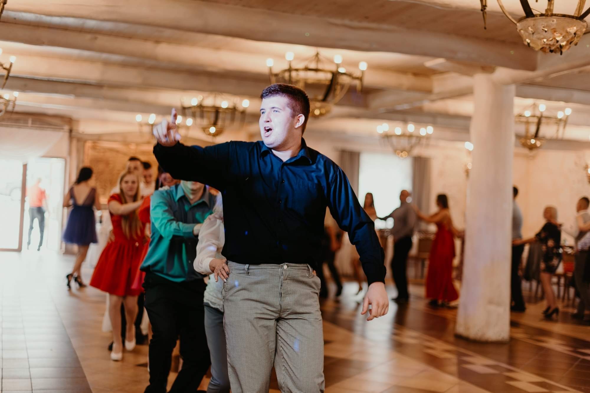 Mężczyzna w czarnej koszuli i spodniach w kratę gestykuluje podczas zabawy tanecznej w przestronnej sali z parkietem i ozdobnymi żyrandolami.