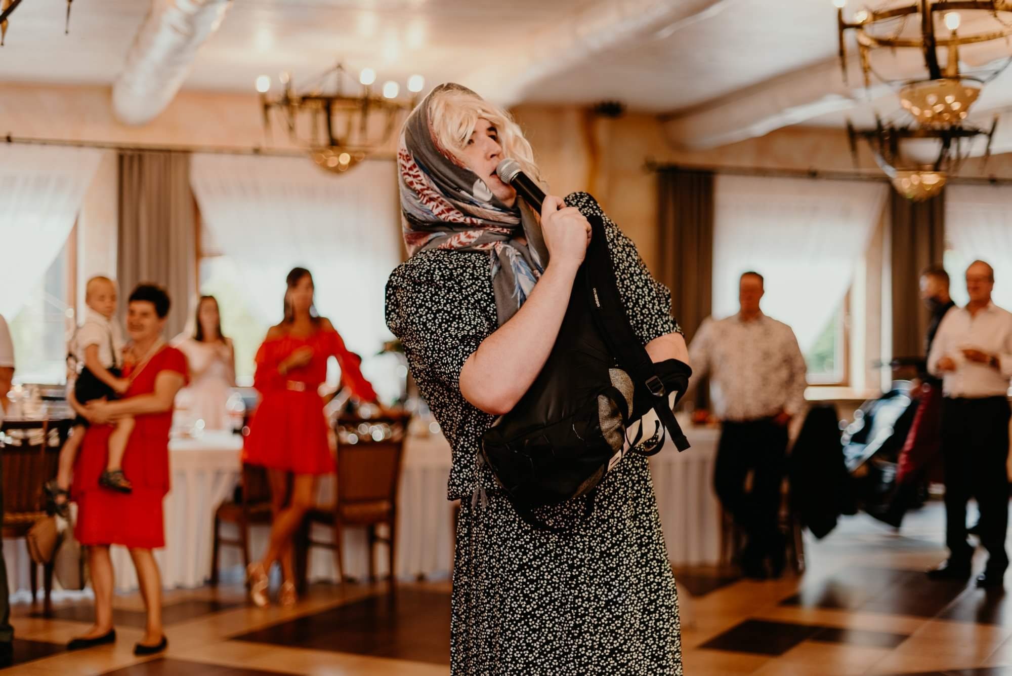 Osoba w przebraniu starszej kobiety, z blond peruką i chustą na głowie, śpiewa do mikrofonu podczas imprezy w sali bankietowej. Na ramieniu ma czarny plecak.