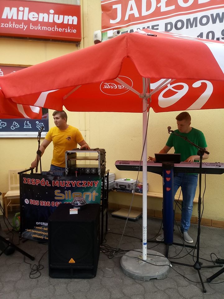 Zespół muzyczny grający na zewnątrz pod czerwonym parasolem Coca-Cola, jeden muzyk obsługuje mikser, drugi gra na keyboardzie Yamaha, widoczne logo zespołu 'Silent' oraz reklama zakładów...