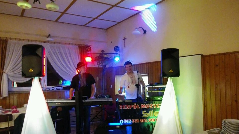 Dwaj mężczyźni w strojach ludowych grający na keyboardzie i obsługujący sprzęt nagłośnieniowy podczas imprezy w sali z drewnianą boazerią i oświetleniem LED.