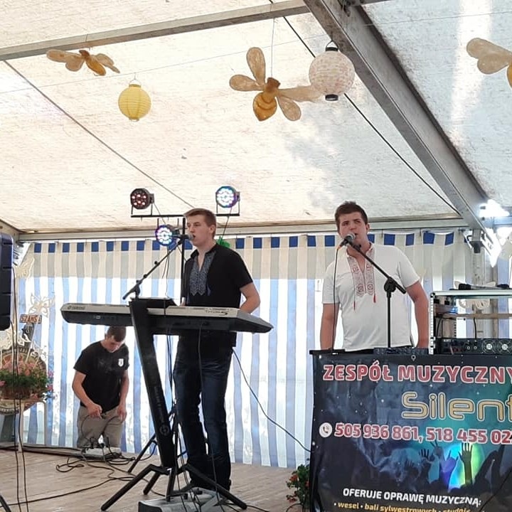 Występujący na scenie dwaj wokaliści w koszulach z haftem, jeden gra na keyboardzie, w tle dekoracje w postaci lampionów i baner reklamowy zespołu.