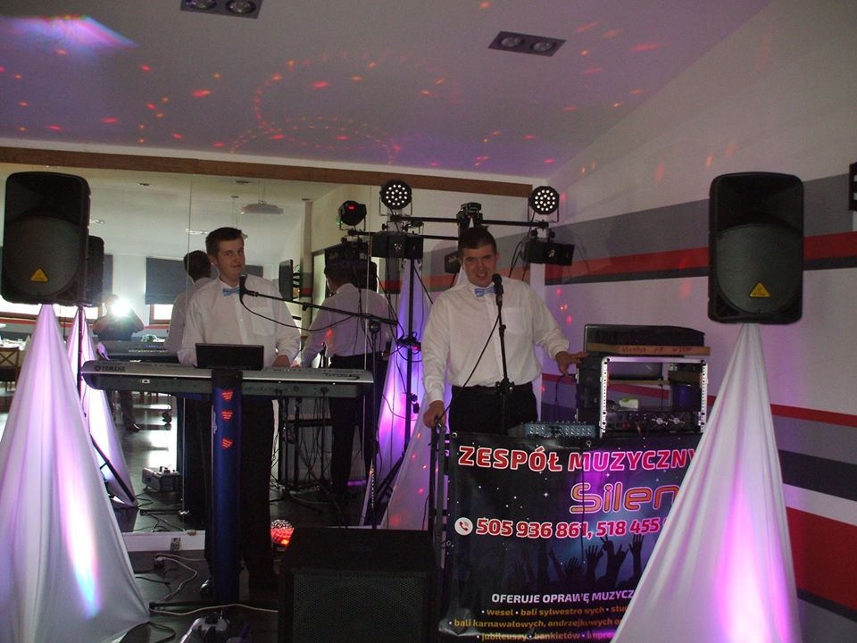 Zespół muzyczny w trakcie występu na sali bankietowej, widoczne instrumenty, nagłośnienie, oświetlenie i baner reklamowy z numerami telefonów.