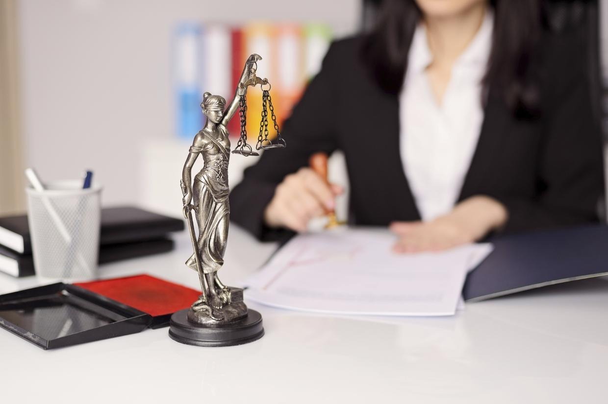 Statuetka Temidy z wagą sprawiedliwości na biurku, w tle kobieta w czarnym żakiecie stempluje dokumenty.