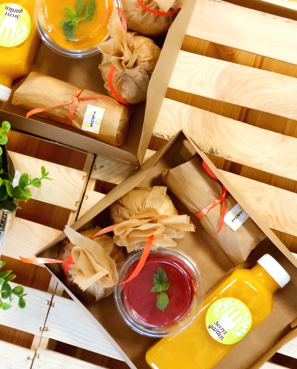 Zestaw śniadaniowy w kartonowych pudełkach na drewnianej palecie: miniburgery z szarpaną wołowiną zawinięte w papier, tortilla z wędzonym łososiem w papierowej owijce z naklejką 'Kurczak', smoothie...