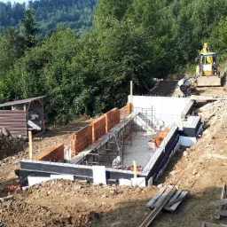 Budowa fundamentów domu na zboczu, widoczne zbrojenia, izolacja pionowa, częściowo wymurowane ściany, betoniarka i koparka w tle na tle zalesionego wzgórza.