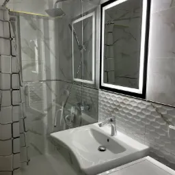 Nowoczesna łazienka z kabiną prysznicową typu walk-in, umywalką nablatową i podświetlanym lustrem, wykończona marmurowymi płytkami i dekoracyjnym pasem z trójwymiarowych płytek.