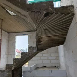 Surowe, betonowe schody spiralne w trakcie budowy domu z bloczków, widoczne zbrojenie i szalunki, mokra podłoga.