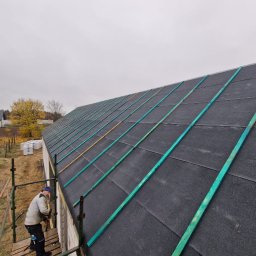 Usługi budowlane Mateusz Schröder - Pracownik na rusztowaniu montuje kontrłaty w kolorze turkusowym na czarnej membranie dachowej nowego domu, widok z góry na dach i okoliczne domy w jesiennej scenerii.