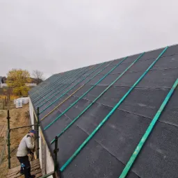 Pracownik na rusztowaniu montuje kontrłaty w kolorze turkusowym na czarnej membranie dachowej nowego domu, widok z góry na dach i okoliczne domy w jesiennej scenerii.