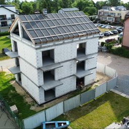 Usługi budowlane Mateusz Schröder - Surowy, trzypiętrowy budynek z betonu komórkowego w trakcie budowy, z widoczną konstrukcją dachu i tymczasowym ogrodzeniem. Ujęcie z góry, otoczenie stanowią inne budynki i zieleń.