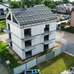 Surowy, trzypiętrowy budynek z betonu komórkowego w trakcie budowy, z widoczną konstrukcją dachu i tymczasowym ogrodzeniem. Ujęcie z góry, otoczenie stanowią inne budynki i zieleń.