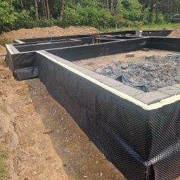 Usługi budowlane Mateusz Schröder - Zabezpieczone fundamenty domu z hydroizolacją i folią kubełkową na tle zieleni i pryzmy piachu. Widoczne zbrojenia i ściany fundamentowe.