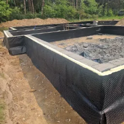 Zabezpieczone fundamenty domu z hydroizolacją i folią kubełkową na tle zieleni i pryzmy piachu. Widoczne zbrojenia i ściany fundamentowe.