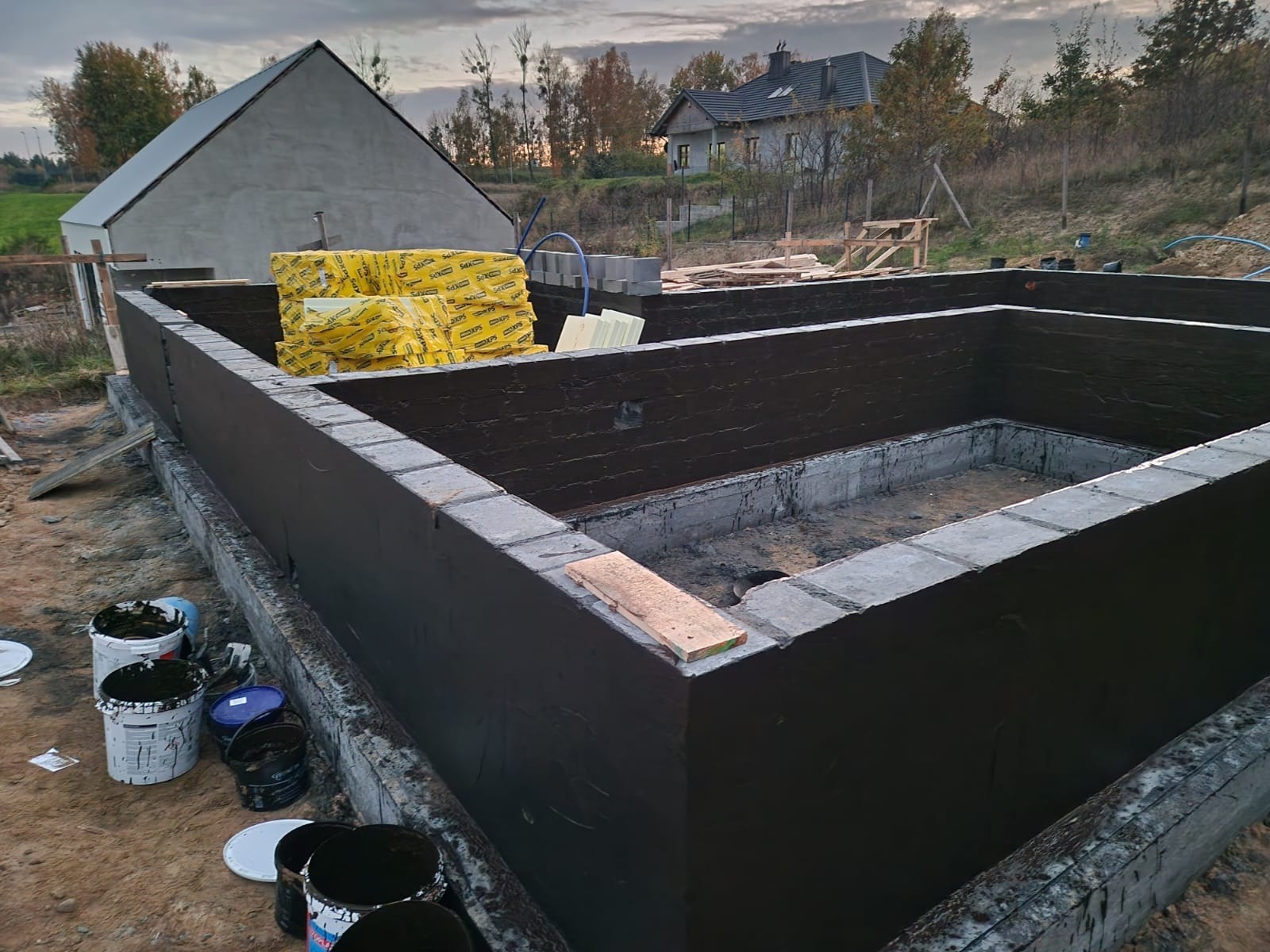 Wykop i murowane ściany fundamentowe zabezpieczone czarną izolacją bitumiczną, na działce budowlanej z widocznymi materiałami izolacyjnymi, pustakami i farbami w wiadrach.