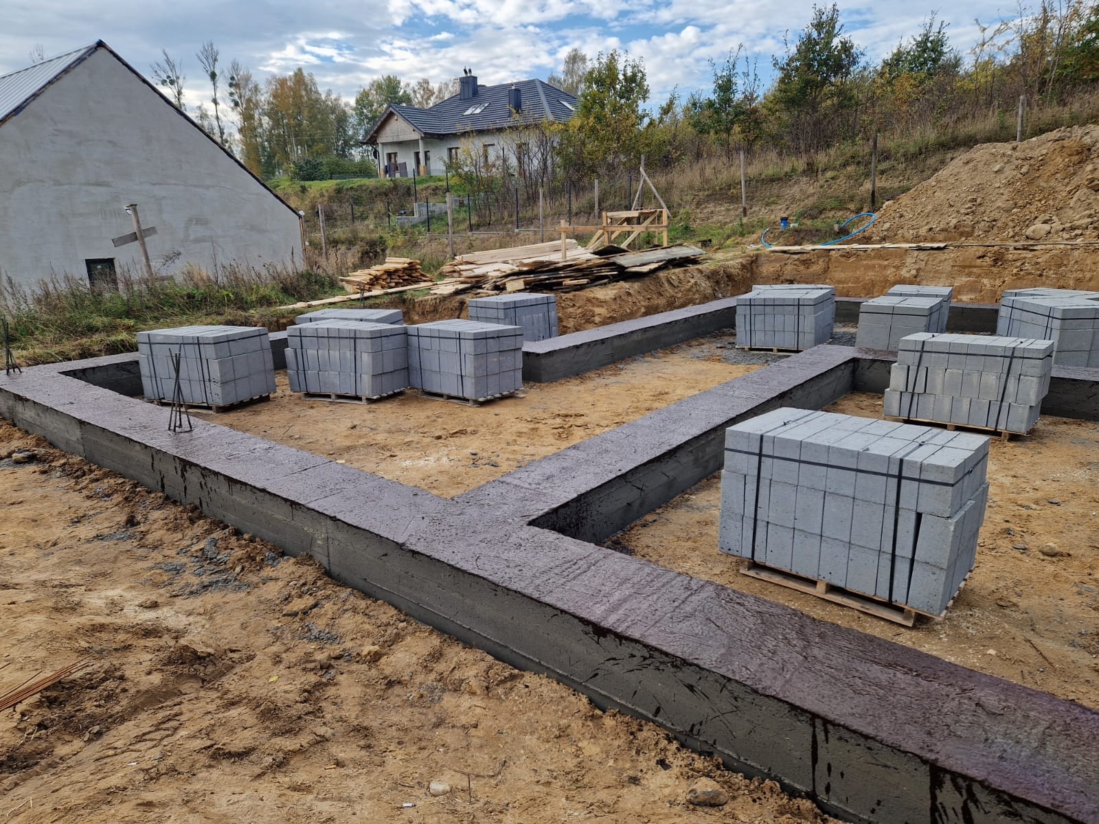 Wylany fundament pod dom jednorodzinny z wystającymi prętami zbrojeniowymi, obok ułożone palety z bloczkami betonowymi, w tle widoczne dwa domy w różnym stanie wykończenia.