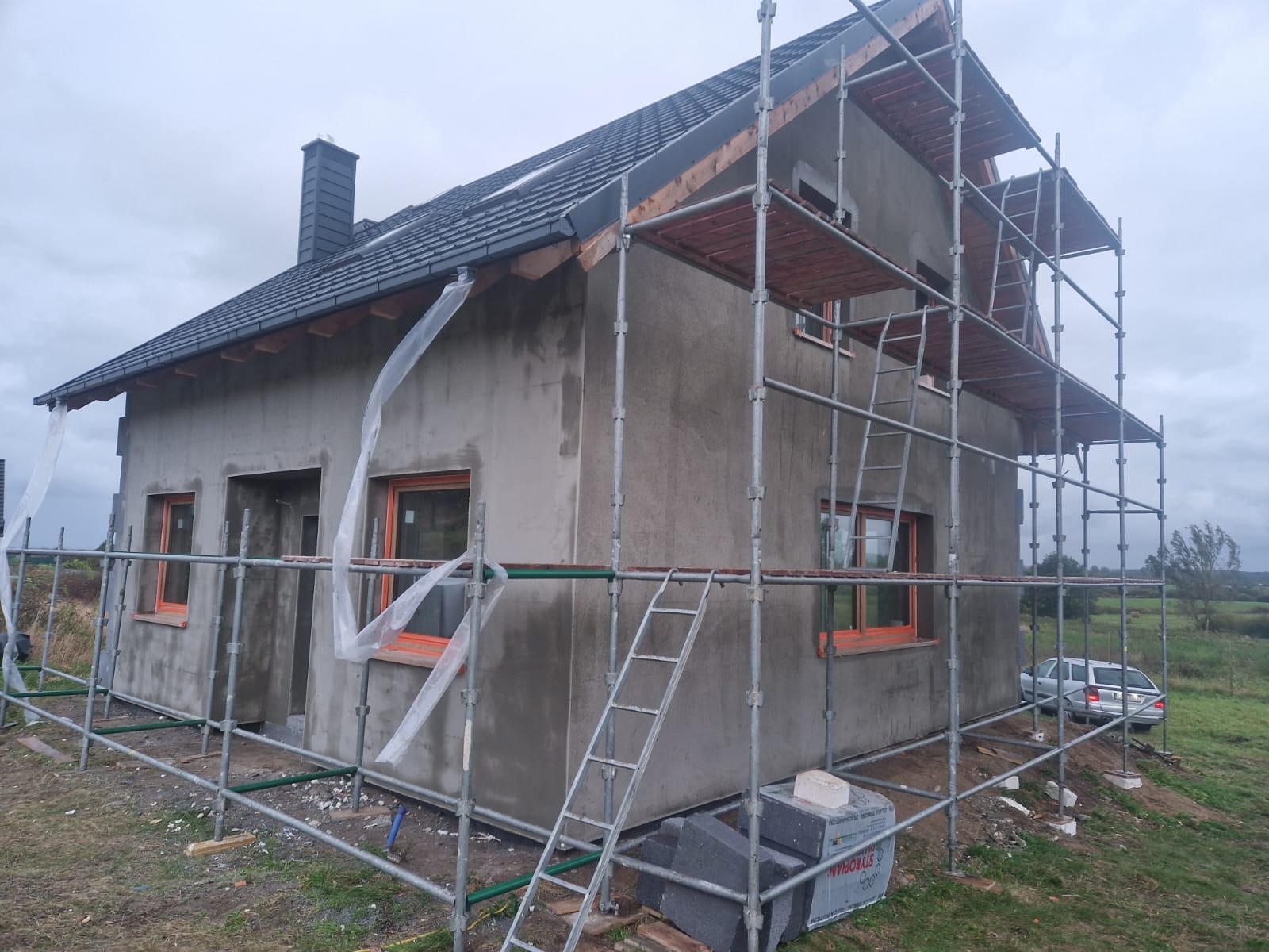 Surowy dom z szarymi ścianami i pomarańczowymi ramami okiennymi otoczony rusztowaniami, widoczne materiały budowlane i samochód w tle