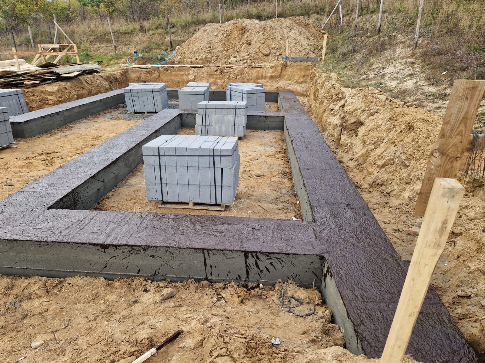 Wykop pod fundamenty z zalewanymi ławami fundamentowymi zabezpieczonymi izolacją bitumiczną, obok ułożone palety z bloczkami betonowymi.