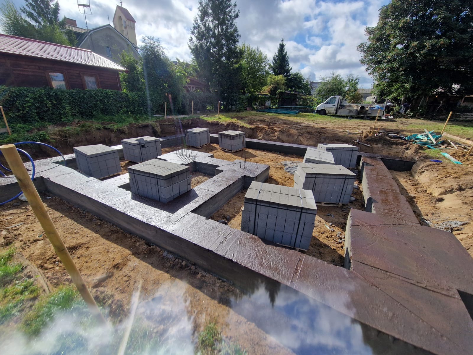 Wylane fundamenty z ułożonymi bloczkami betonowymi na paletach, widok z góry na plac budowy z budynkiem i drzewami w tle, widać świeżo wylany beton.