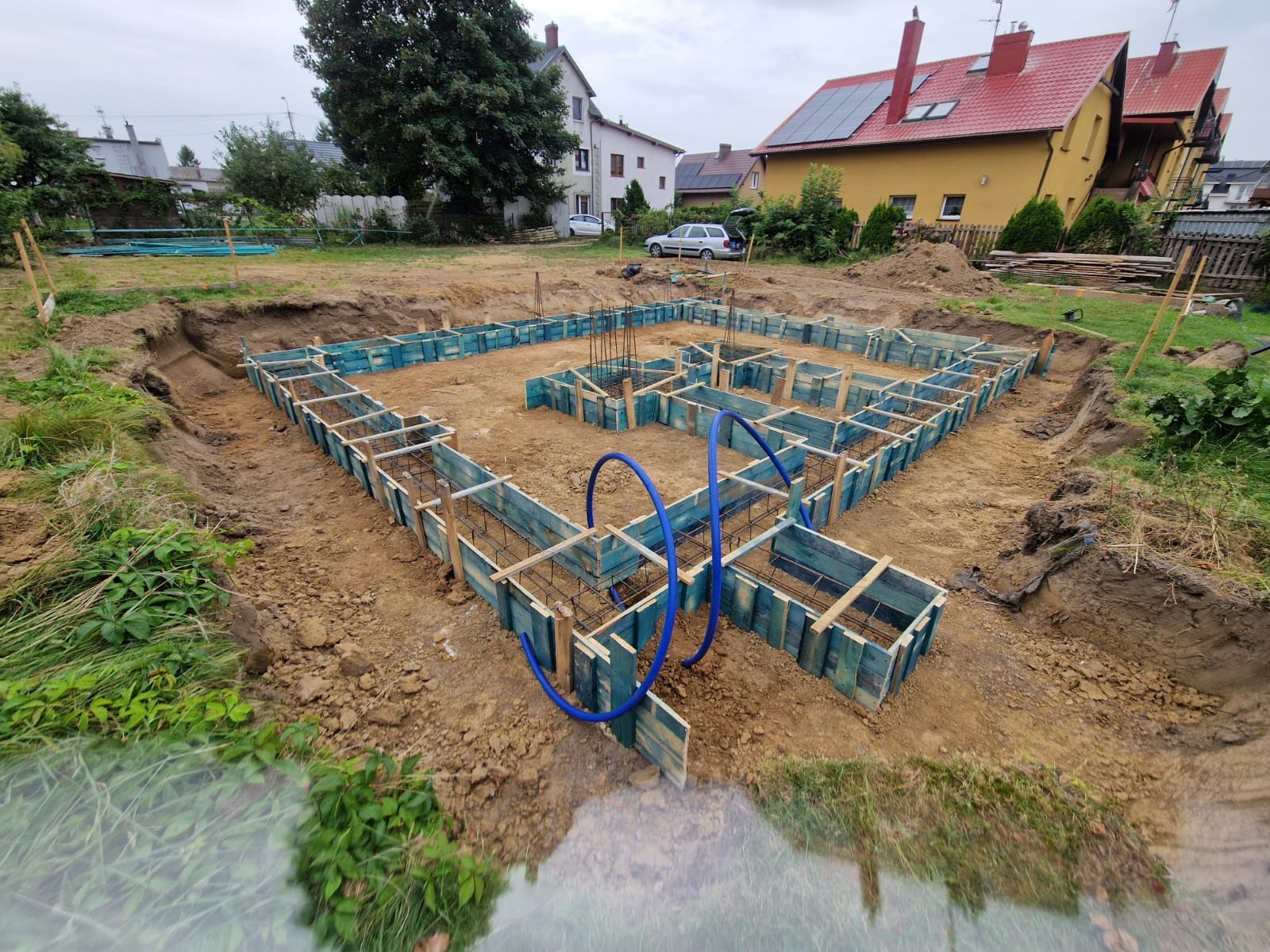 Przygotowanie do wylewki fundamentów: szalunki z desek pomalowanych na niebiesko, zbrojenie stalowe, wykop ziemny, w tle dom z czerwonym dachem i panelami słonecznymi.