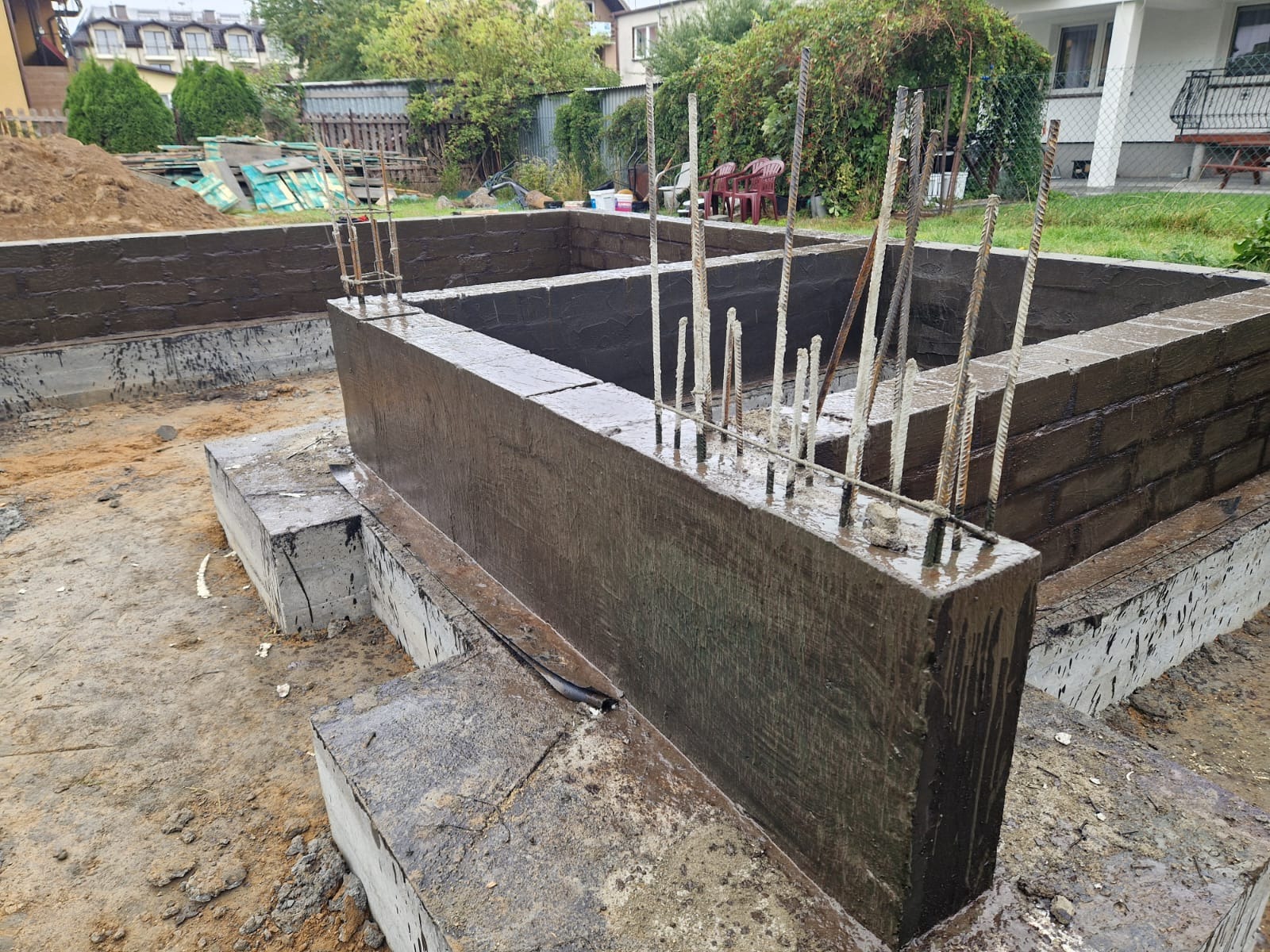 Wykopany fundament z wystającymi prętami zbrojeniowymi, zabezpieczony czarną powłoką hydroizolacyjną, widok z bliska.