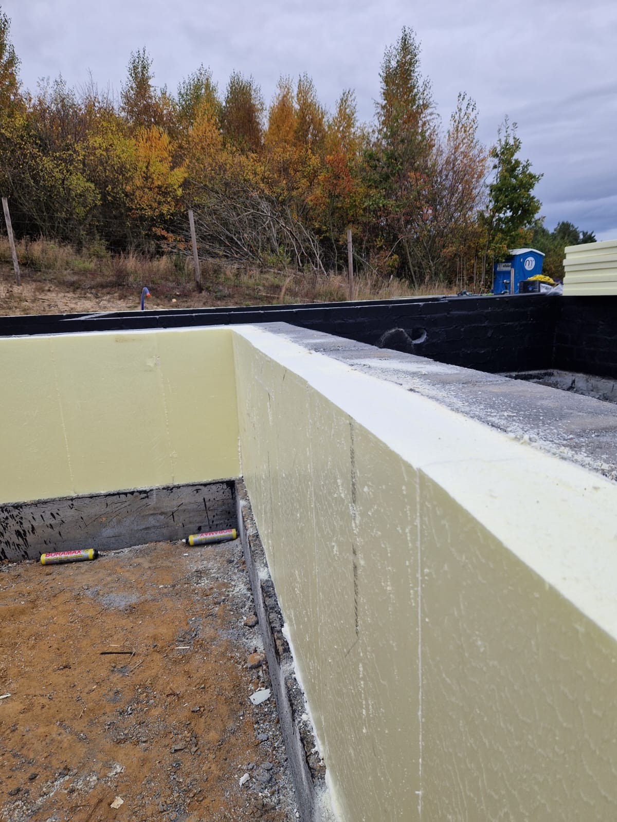 Ocieplone ściany fundamentowe z widocznymi tubami kleju montażowego Duramax, z czarną hydroizolacją i jesiennym krajobrazem w tle.