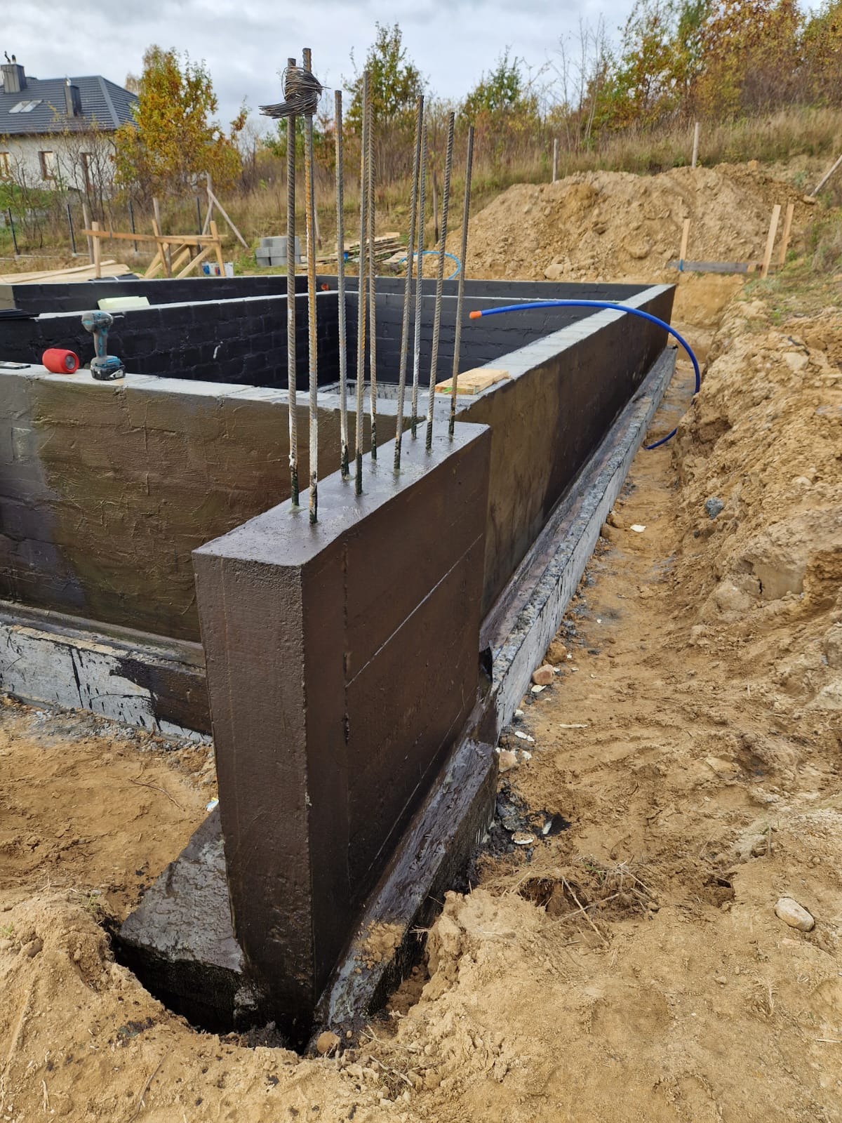 Zabezpieczone hydroizolacją fundamenty domu w trakcie budowy, wystające pręty zbrojeniowe, widoczna czarna, pomalowana ściana fundamentowa oraz narzędzia budowlane w tle.