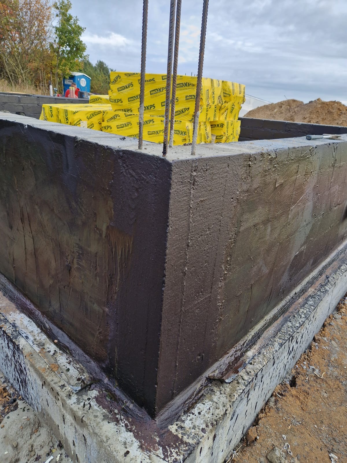 Zabezpieczony hydroizolacją fundament z wystającymi prętami zbrojeniowymi, w tle stos żółtych płyt izolacyjnych Hoch XPS i częściowo widoczny teren budowy.
