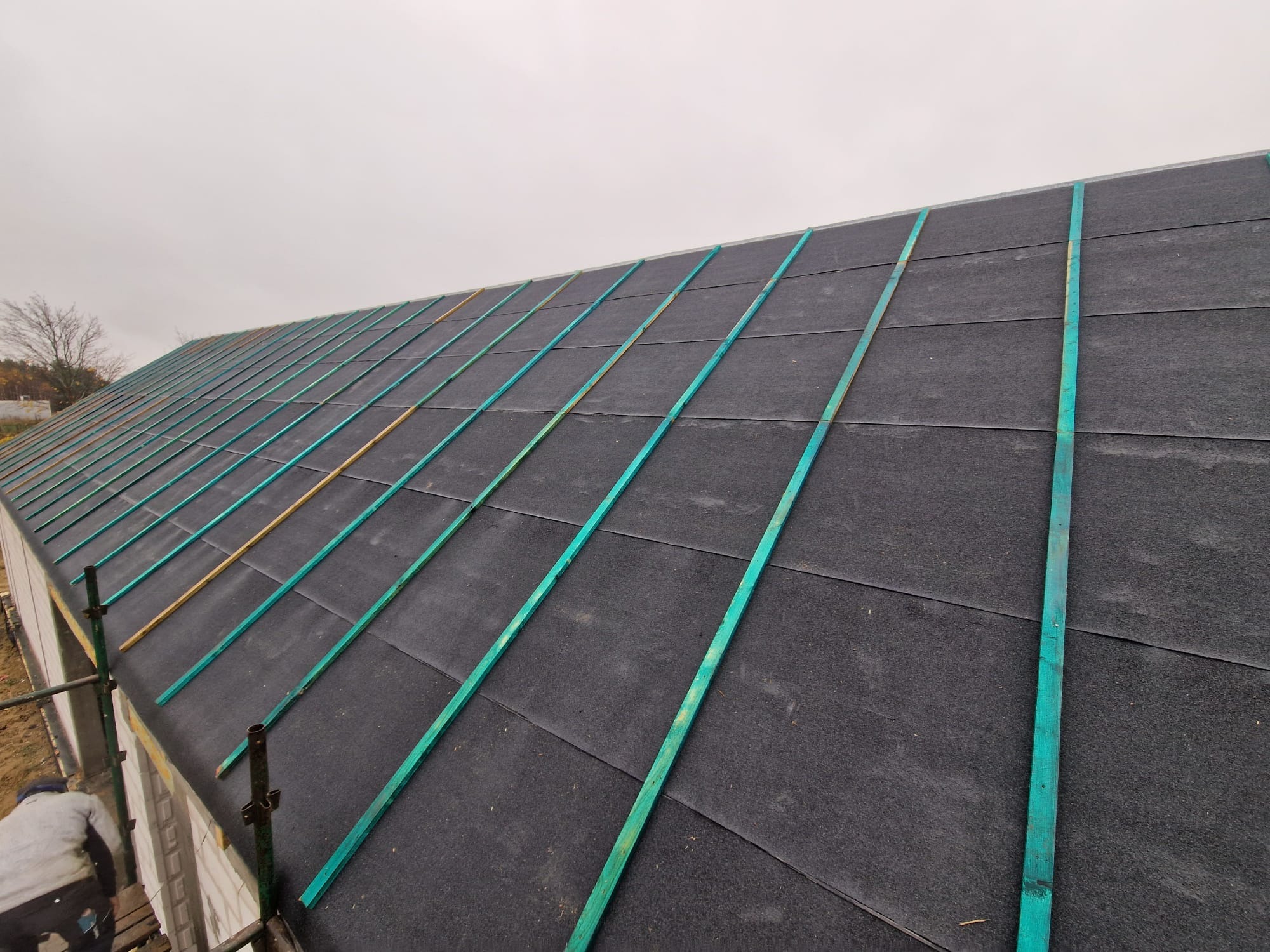 Widok z góry na dach domu w trakcie budowy, pokryty czarną membraną i łatami zaimpregnowanymi na zielono, przygotowany pod pokrycie dachowe.