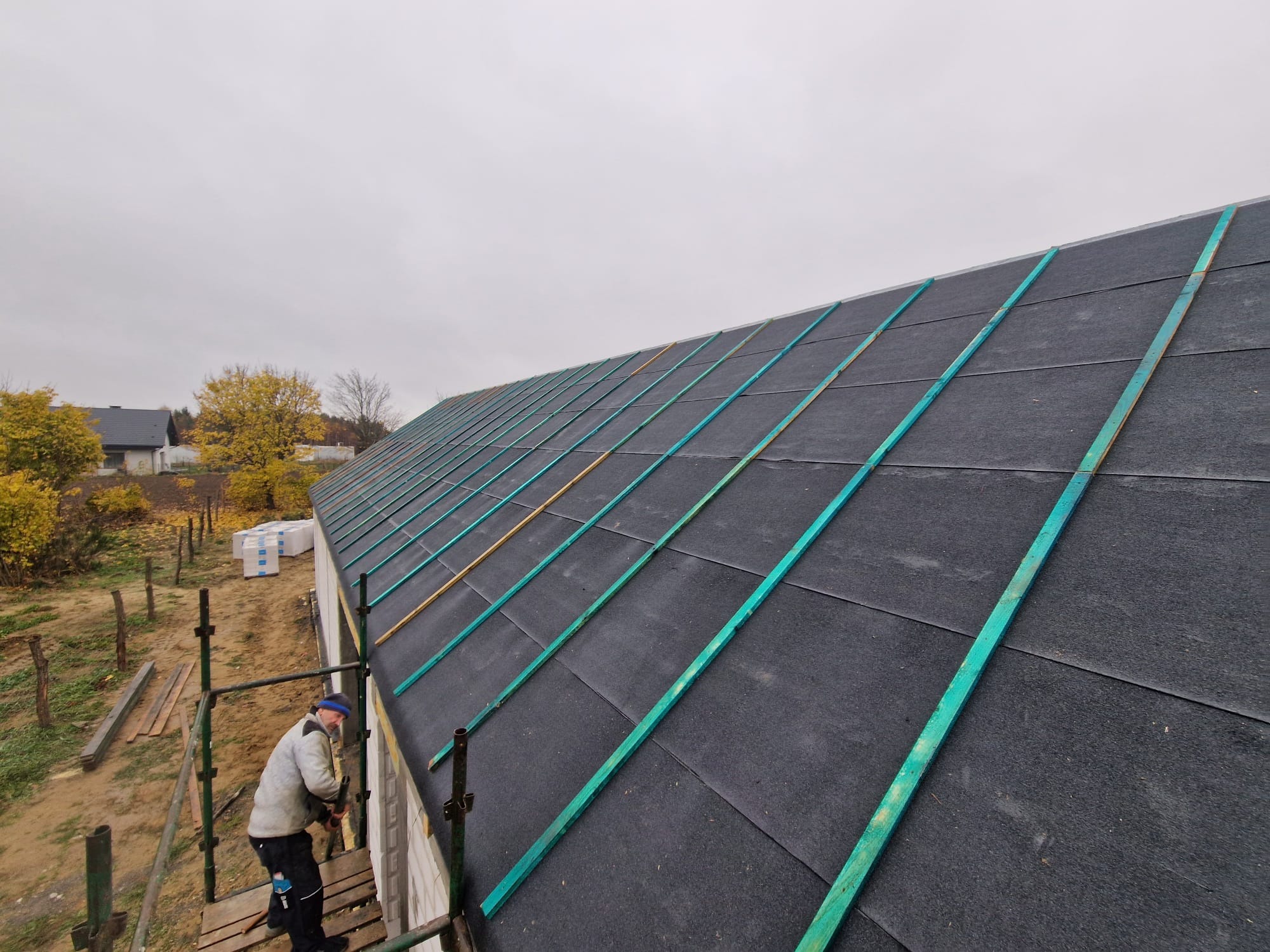 Pracownik na rusztowaniu montuje kontrłaty w kolorze turkusowym na czarnej membranie dachowej nowego domu, widok z góry na dach i okoliczne domy w jesiennej scenerii.