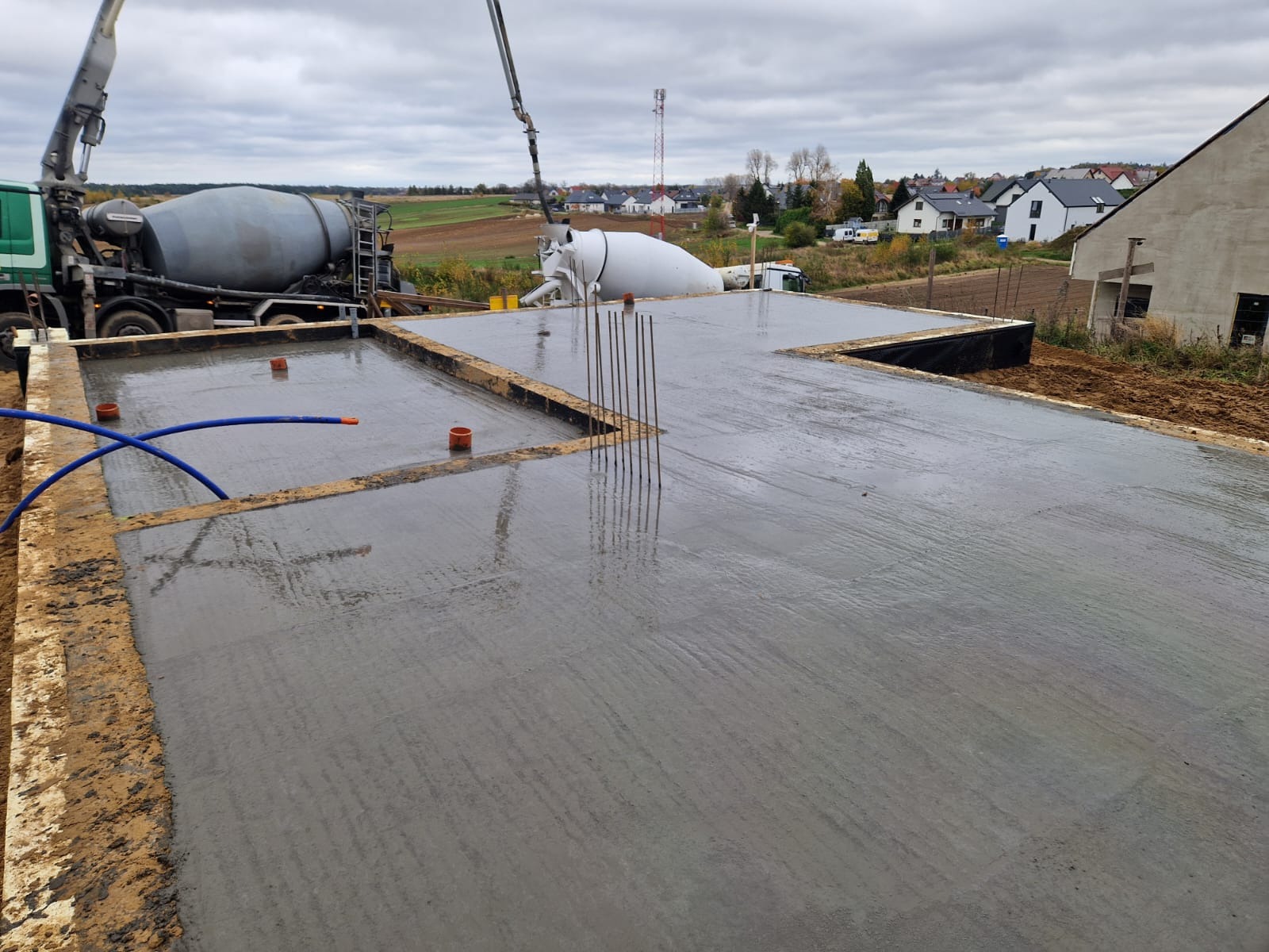 Świeżo wylany, mokry beton na płycie fundamentowej, z widocznymi wystającymi prętami zbrojeniowymi, szalunkami i rurami kanalizacyjnymi, z betoniarką i pompą do betonu w tle na tle krajobrazu...