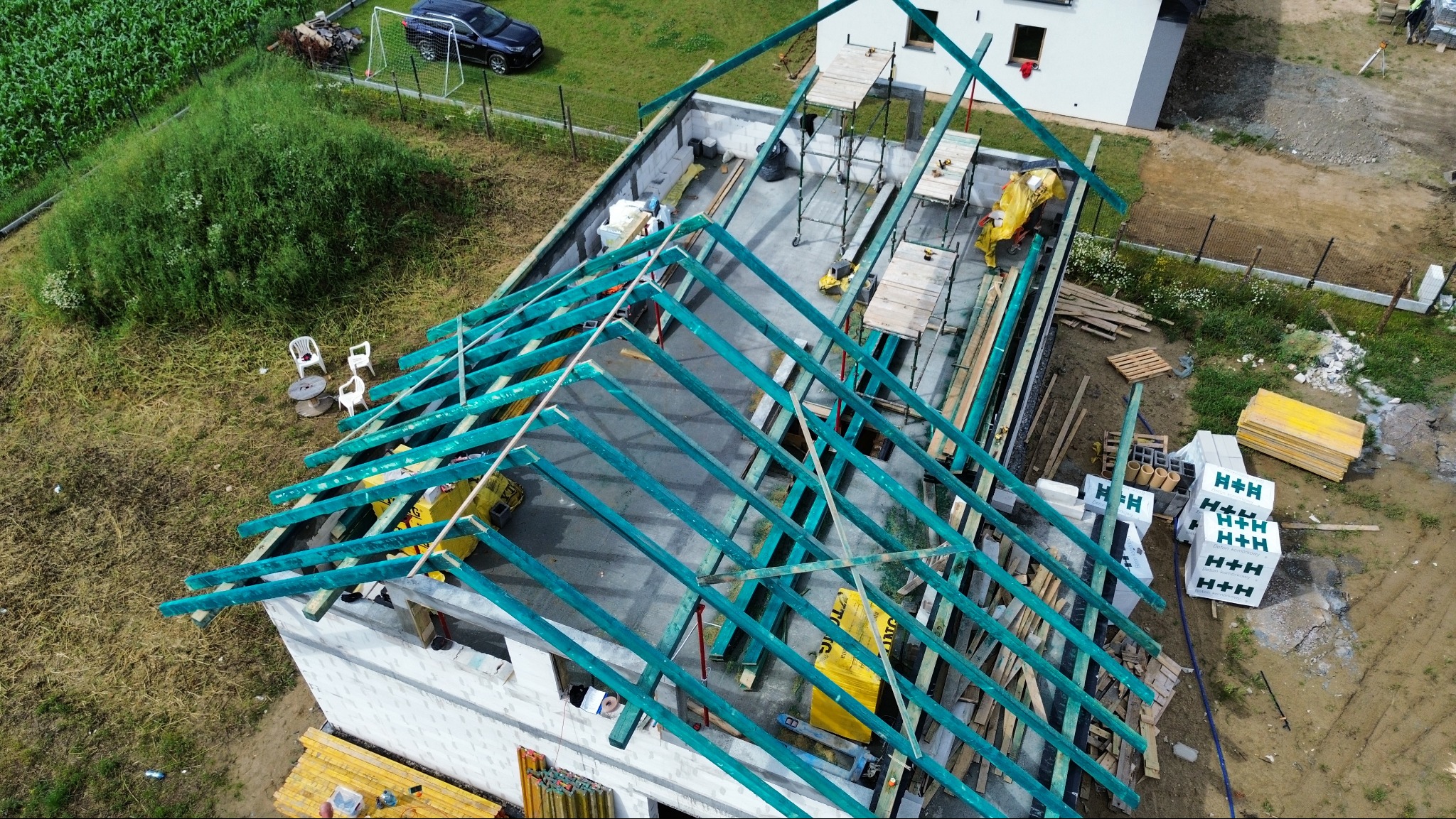 Budowa domu: widok z góry na wznoszone ściany i konstrukcję dachu z zielonych belek. Na działce materiały budowlane i rusztowania. W tle dom sąsiada i auto.