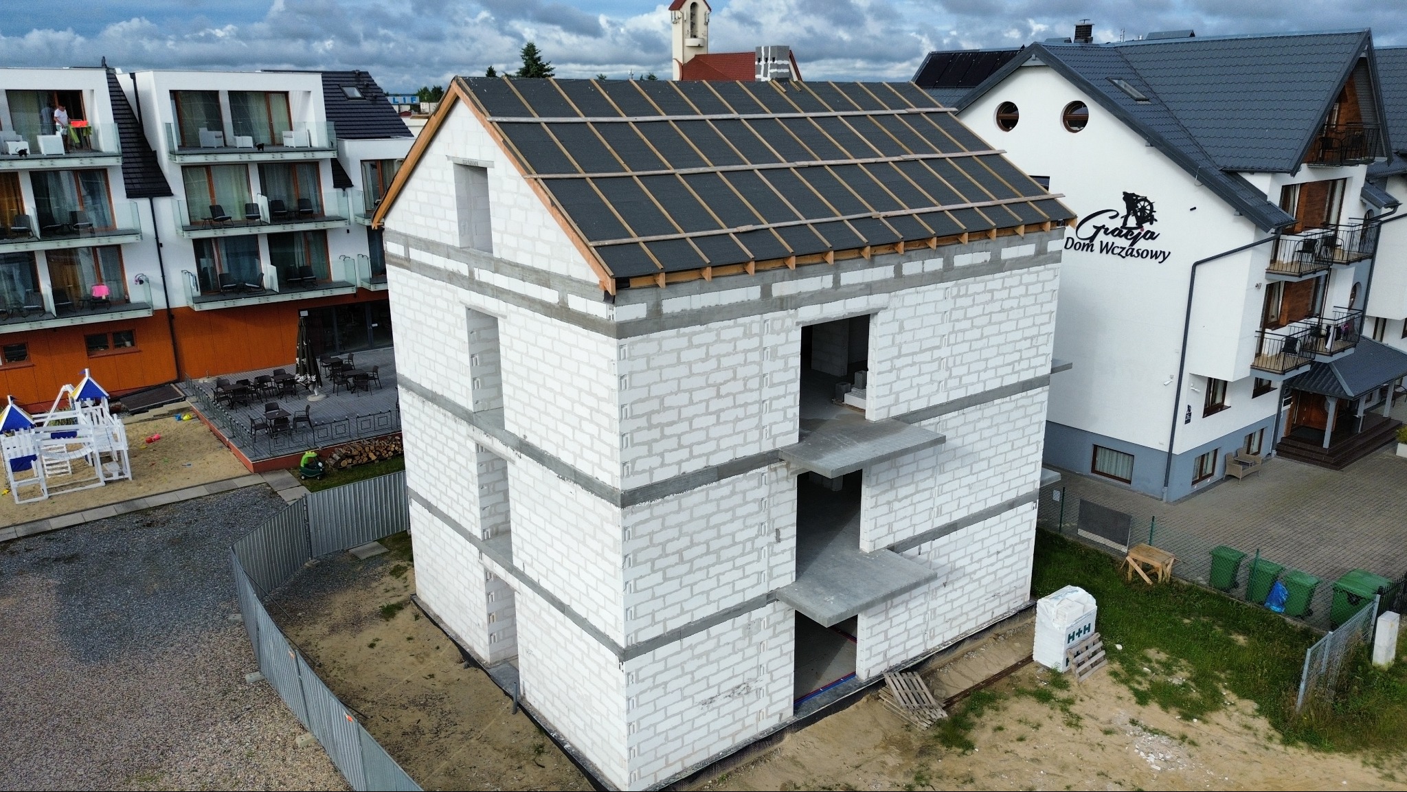 Surowy stan domu z betonu komórkowego, widok z góry. Częściowo zadaszony, obok budynek hotelowy i plac zabaw. Widoczne pustaki H+H na palecie.