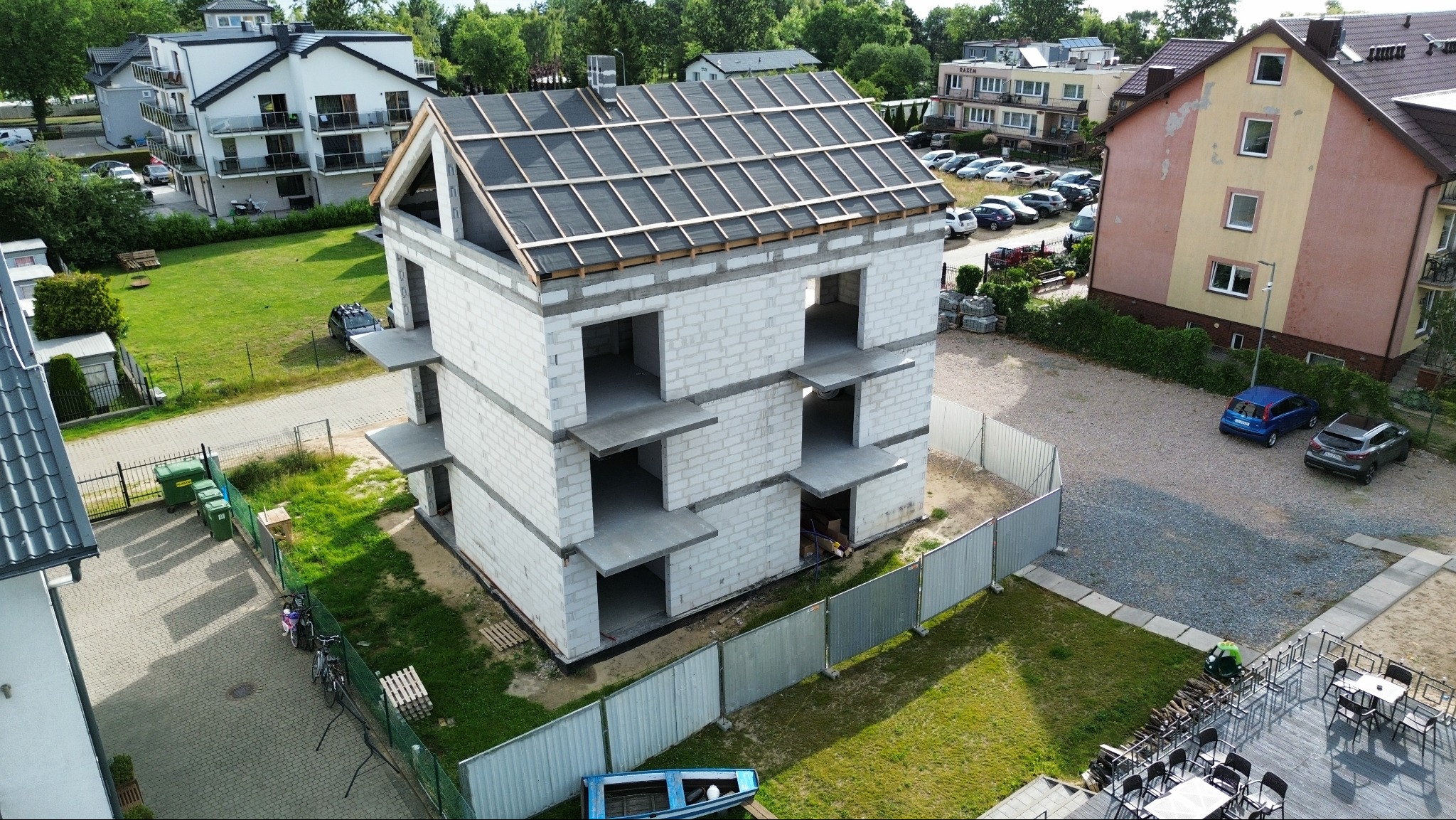 Surowy, trzypiętrowy budynek z betonu komórkowego w trakcie budowy, z widoczną konstrukcją dachu i tymczasowym ogrodzeniem. Ujęcie z góry, otoczenie stanowią inne budynki i zieleń.