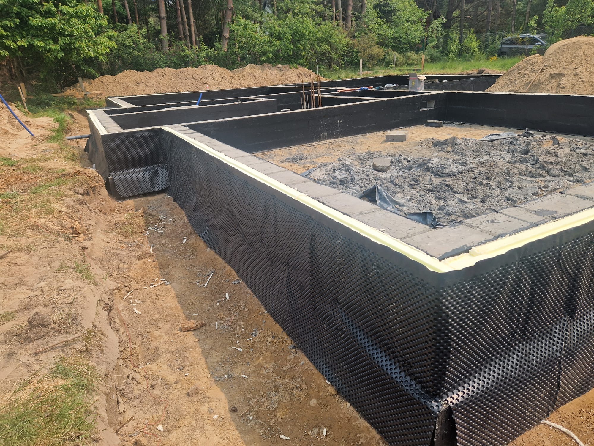 Zabezpieczone fundamenty domu z hydroizolacją i folią kubełkową na tle zieleni i pryzmy piachu. Widoczne zbrojenia i ściany fundamentowe.