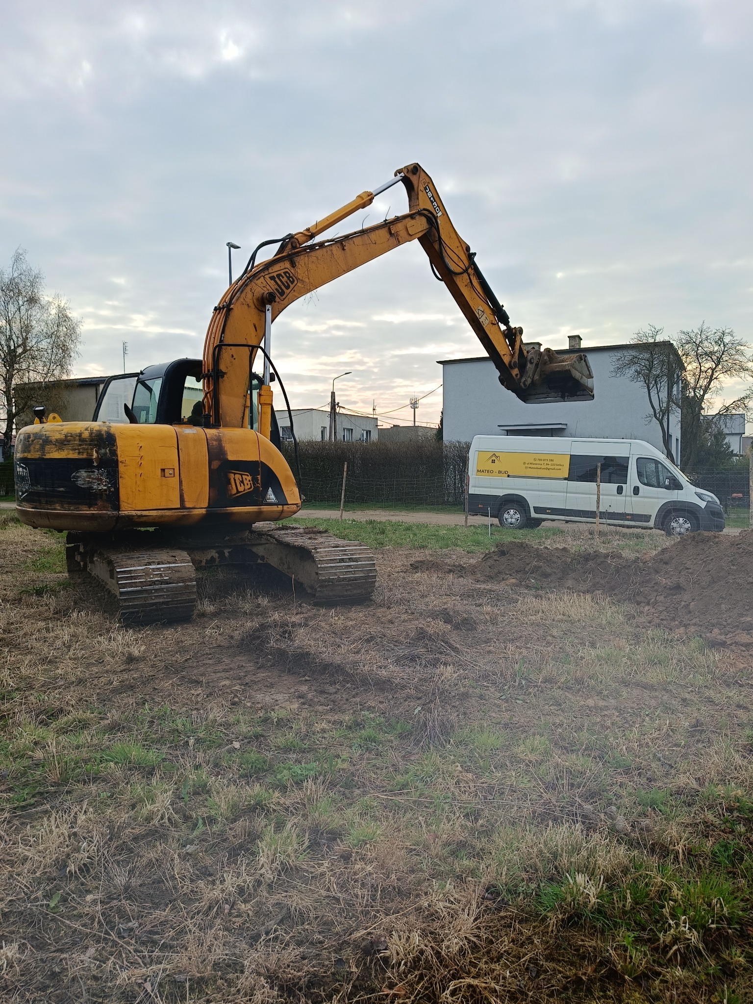 Żółta koparka JCB na placu budowy, obok biały bus z logo firmy budowlanej. Prace ziemne w toku, pochmurne niebo w tle.