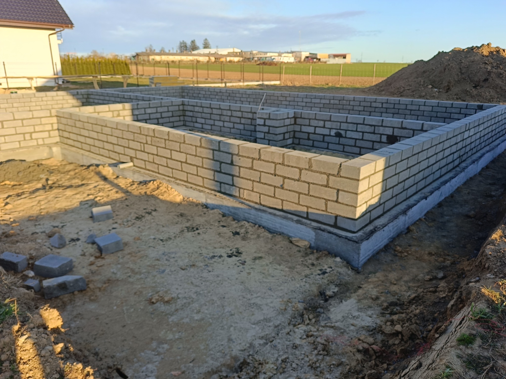 Fundamenty domu z betonu komórkowego, widok z góry na częściowo ukończoną konstrukcję. W tle dom w budowie i sterta ziemi.