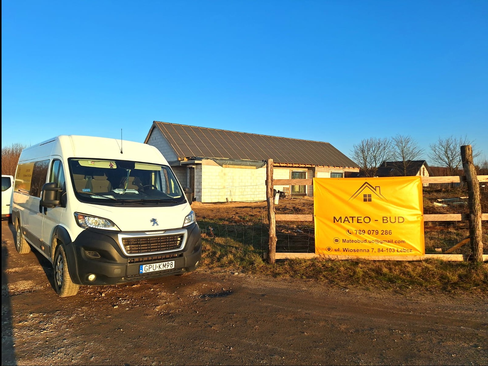 Budowa domu jednorodzinnego w trakcie realizacji, widoczny biały bus firmy budowlanej i baner reklamowy na tle budynku w stanie surowym otwartym.