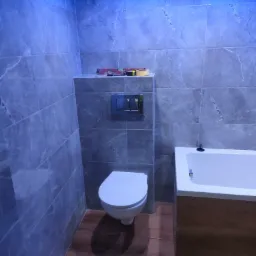 Wykończona łazienka z szarymi płytkami imitującymi marmur, drewnopodobną podłogą, białą wanną i toaletą, oświetleniem LED w suficie, poziomica i taśma miernicza na półce.